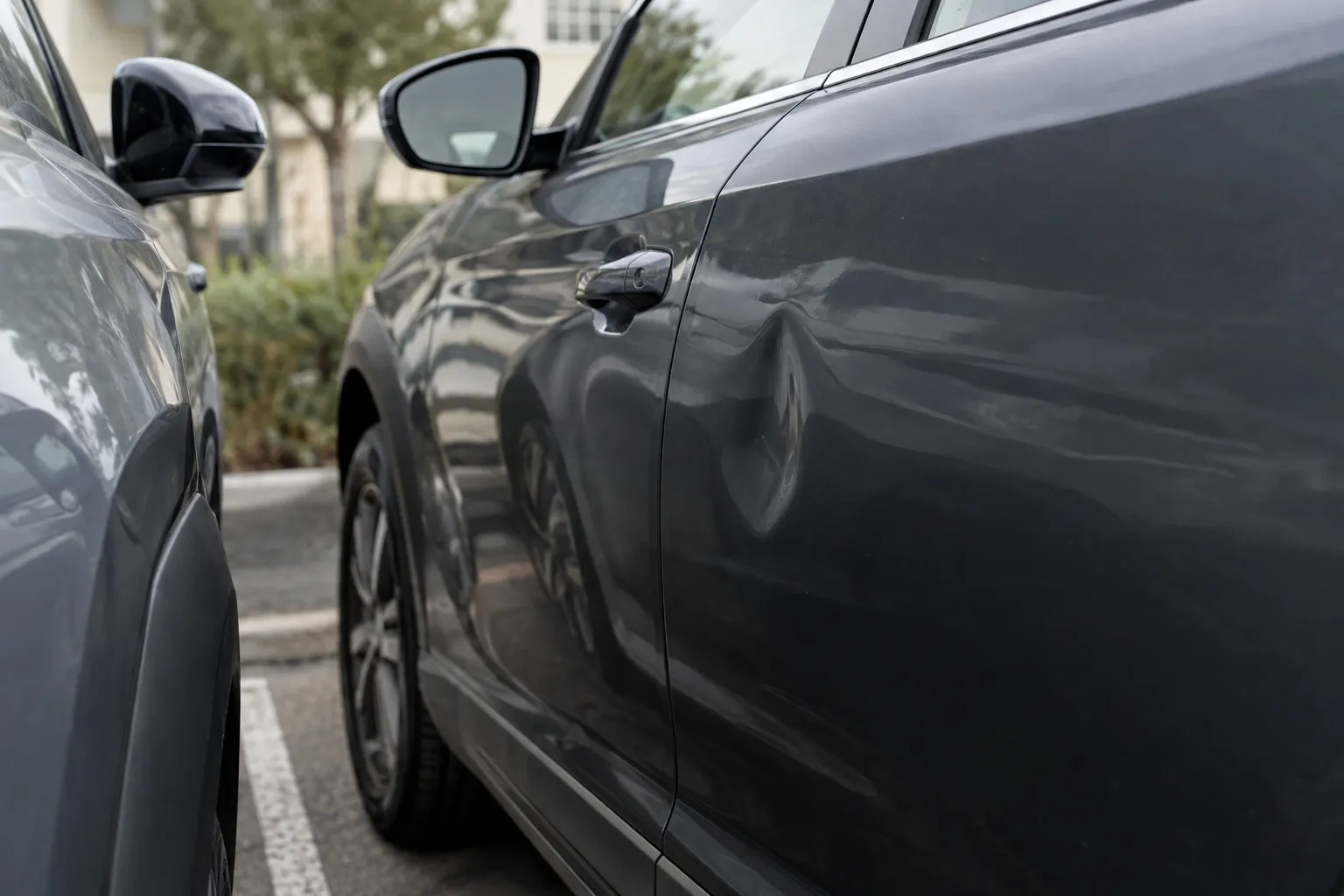 Gros plan sur une bosse sur la portière d'une voiture sombre garée à côté d'une autre, sur un parking de bureaux.